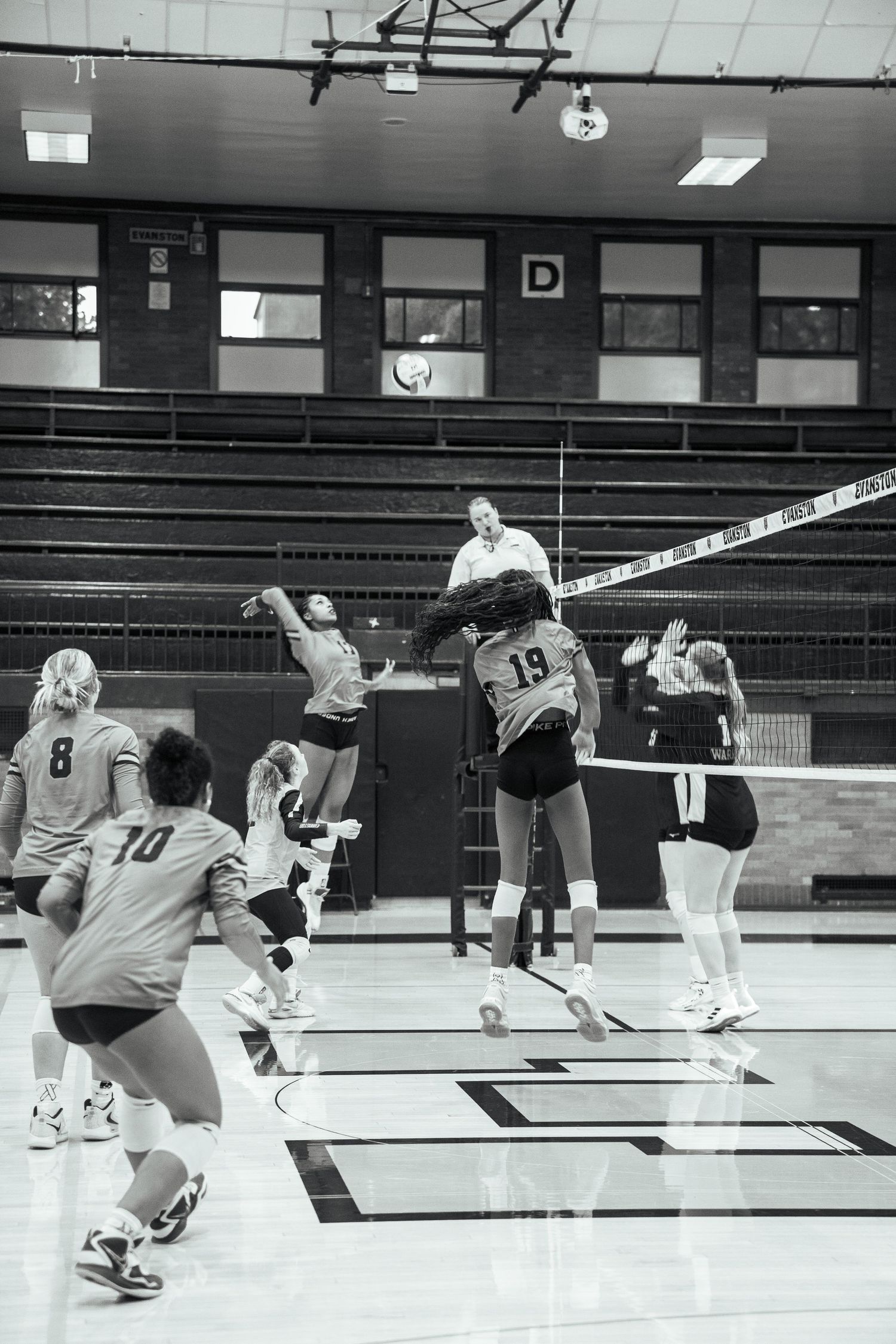 Indoor volleyball match showing players jumping at the net for a block.