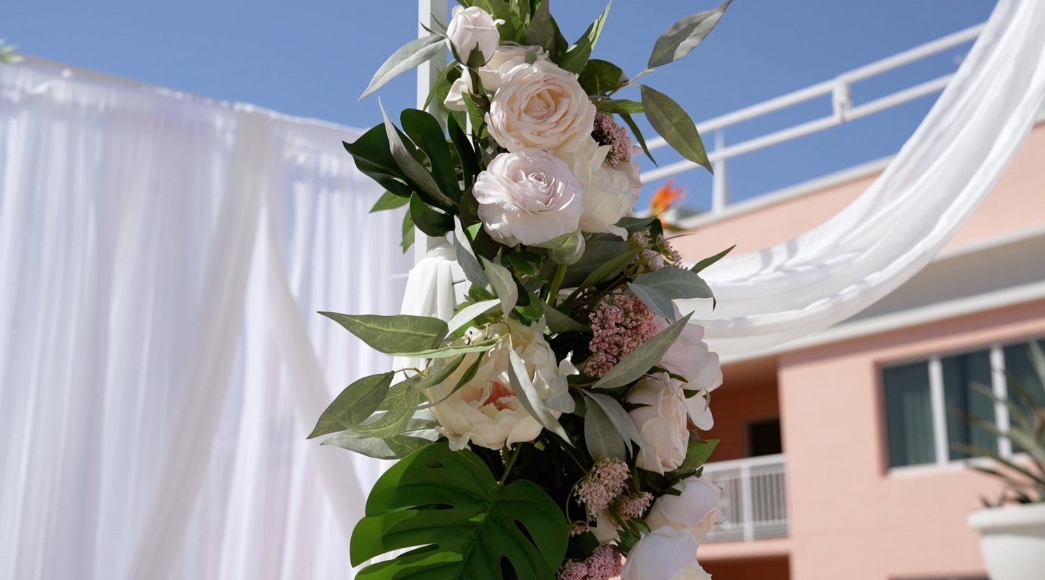White wedding arch decorated with fresh roses and greenery against a pink building.