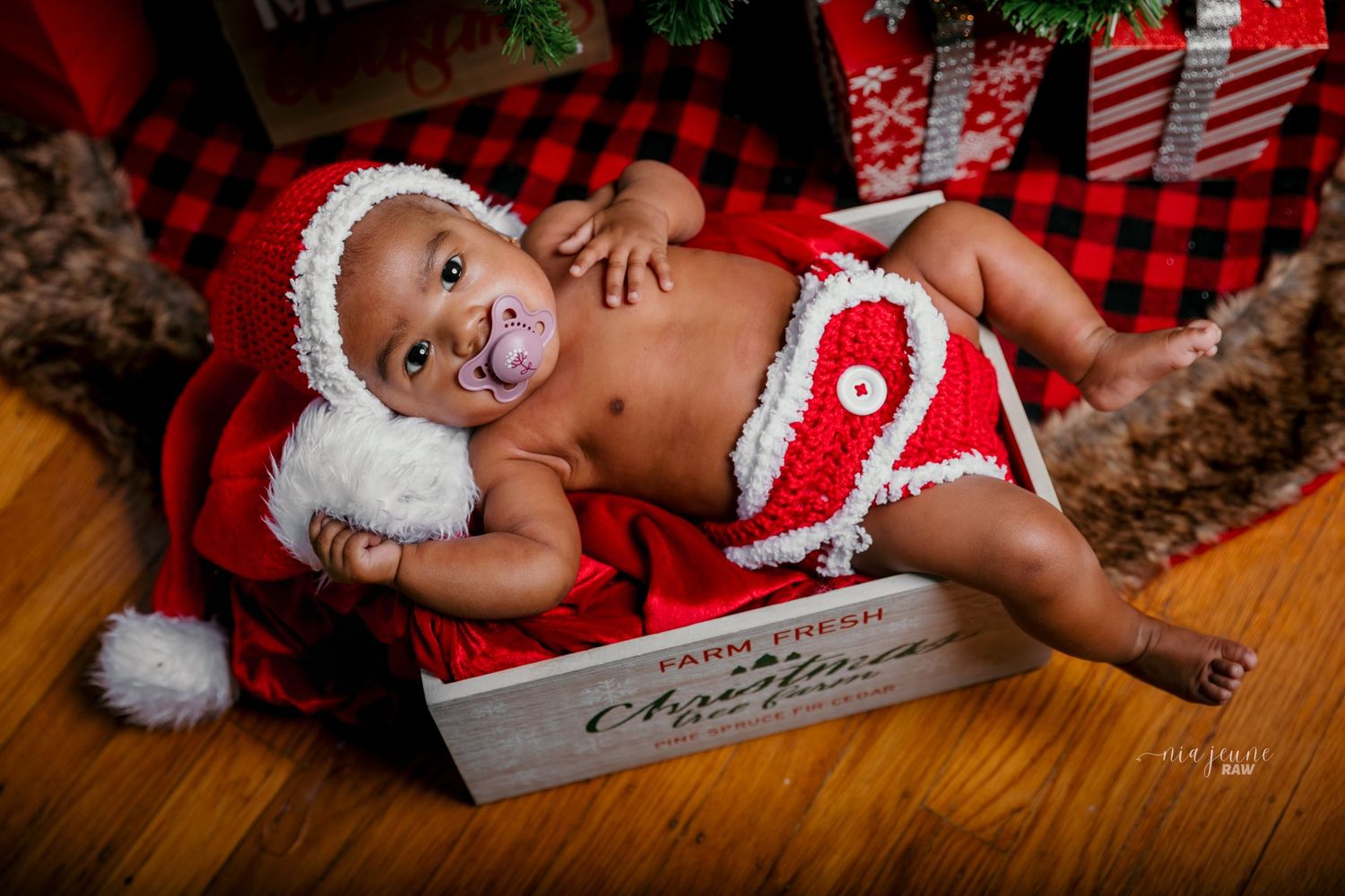 Baby wearing festive Santa outfit lying on plaid blanket among Christmas decorations.