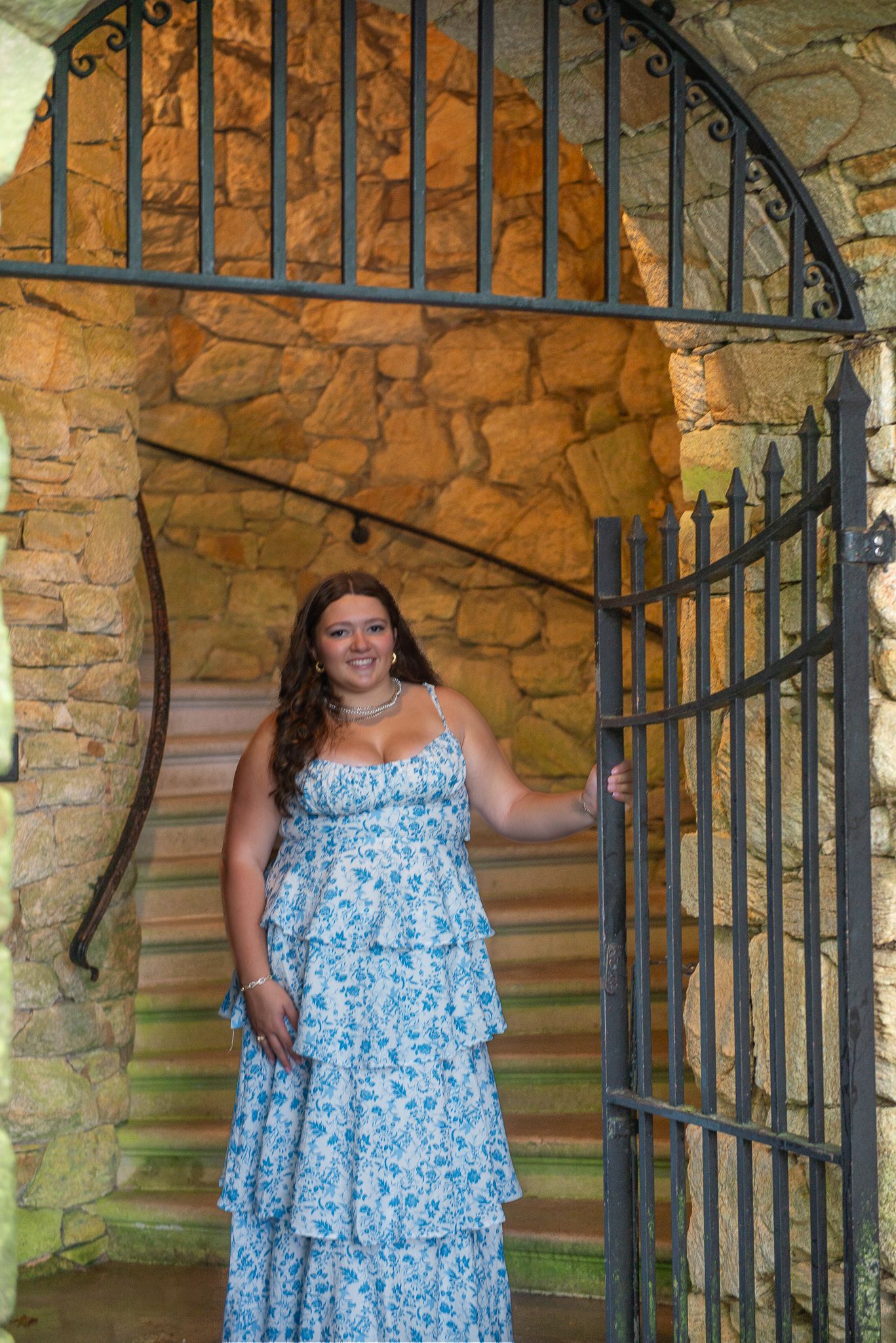 Senior portrait in blue dress standing by stone archway and decorative iron gate.