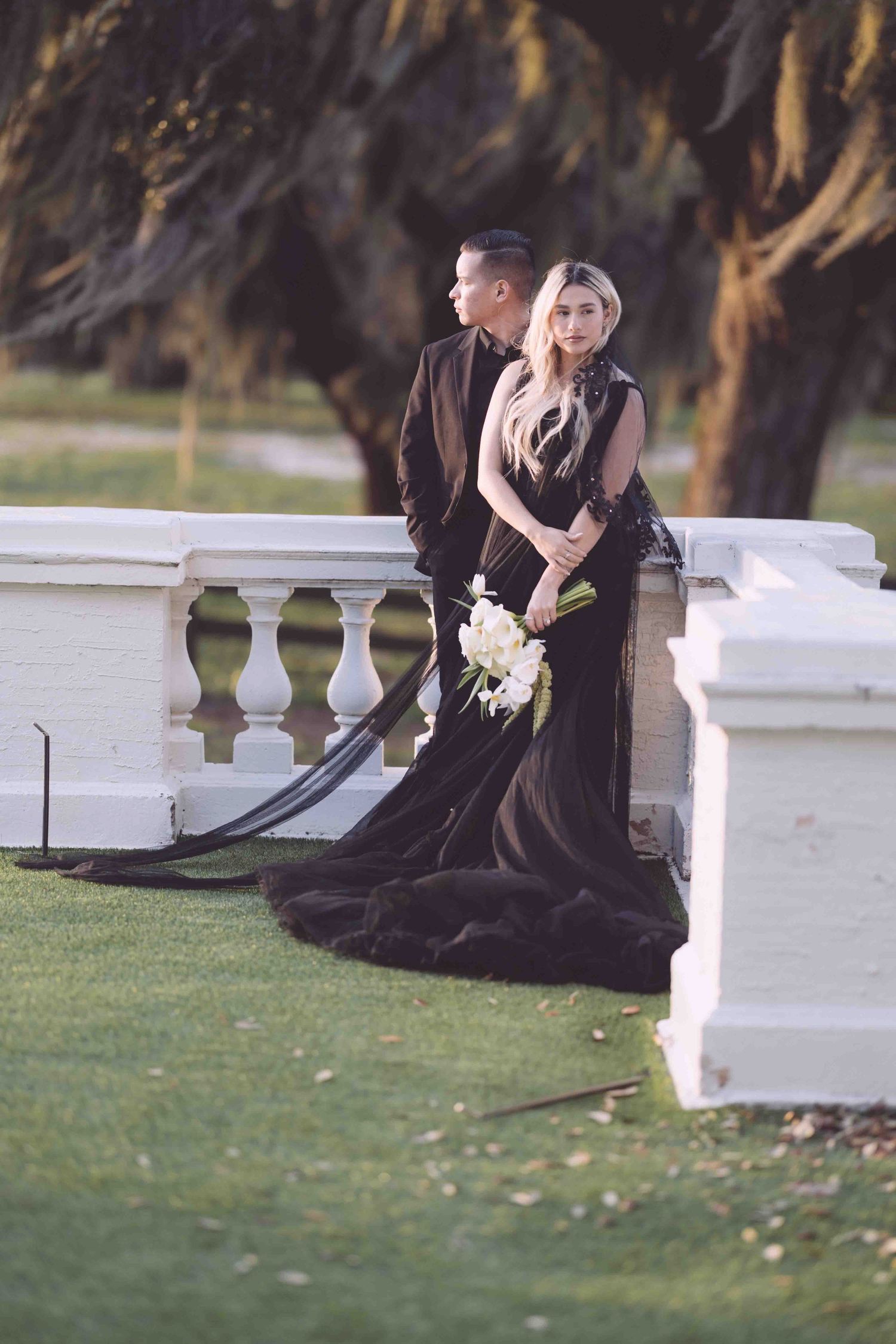 Elegant couple in formal black attire posing by a white balustrade.
