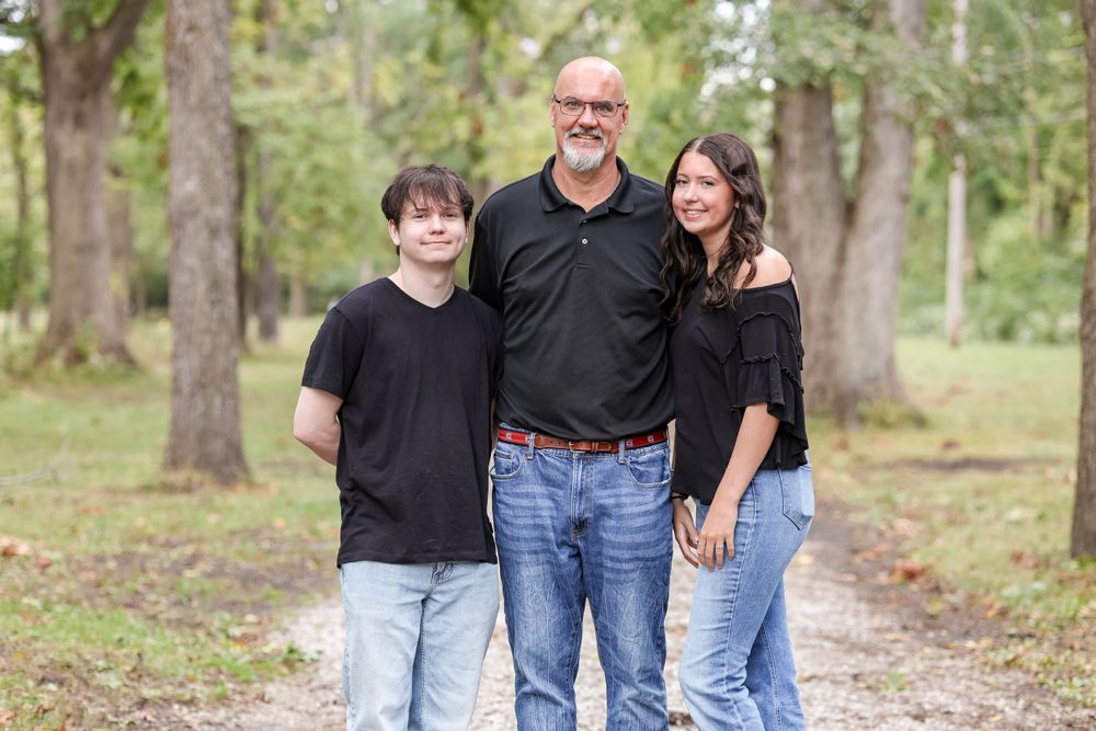 Father takes picture with his two grown kids, all while wearing black, on a tree-lined trail.