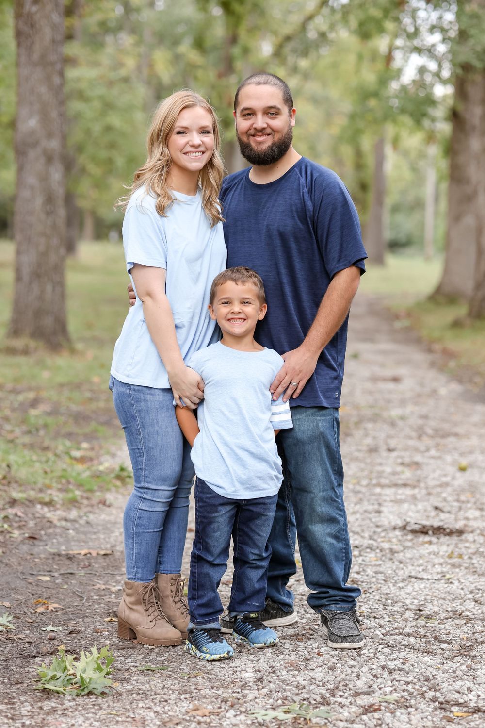 Couple with their son stand smiling on a trail lined by trees with green leaves