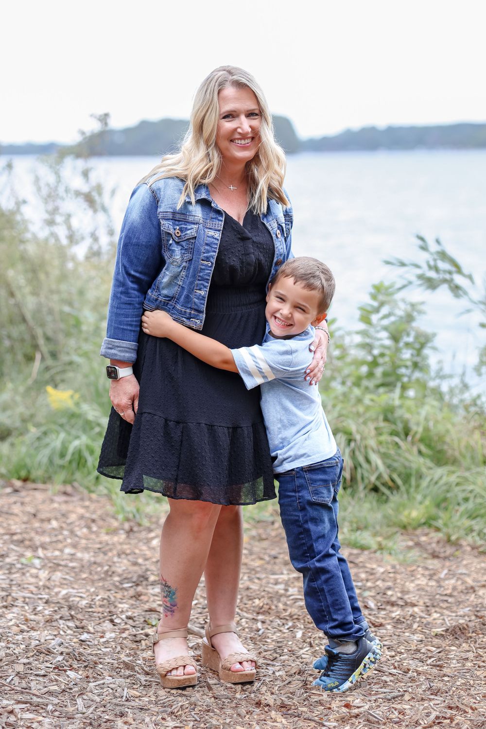 Grandmother and child embrace while standing on a lakeside trail, wearing denim and casual attire.