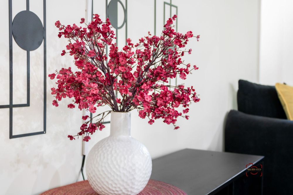 Vibrant red cherry blossom branches arranged in a textured white ceramic vase on a table with dark furniture.