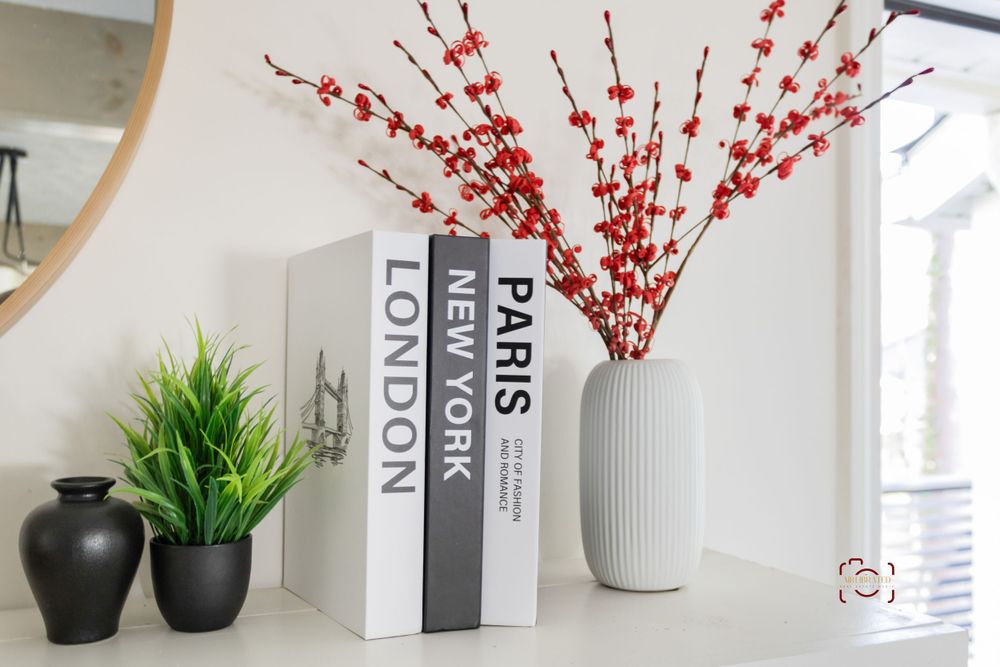 Modern home decor arrangement featuring books, a white vase with red berries, and small potted plants on a shelf.