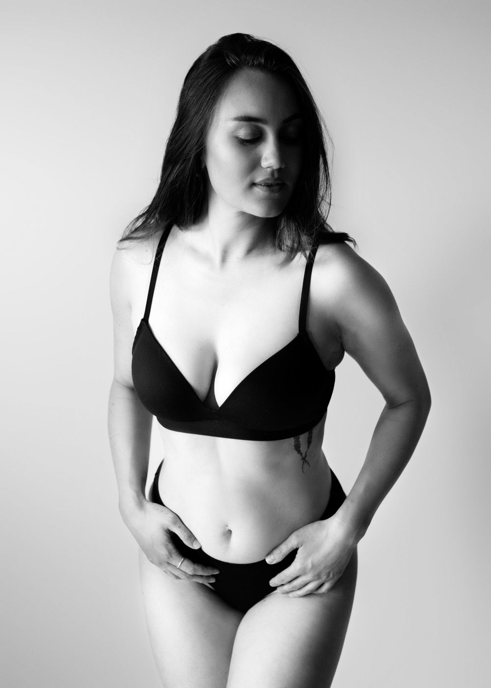 Black and white boudoir portrait of woman in black underwear standing looking off camera.