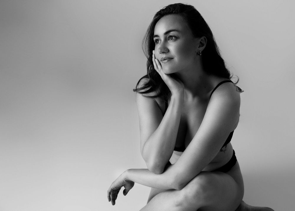 Black and white boudoir portrait of woman sitting on the floor with legs crossed and head in her hand.