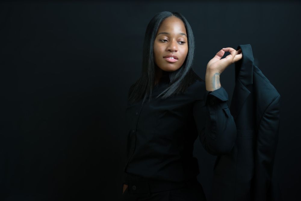 Dramatic portrait against dark background showing figure in black hoodie with long straight hair.