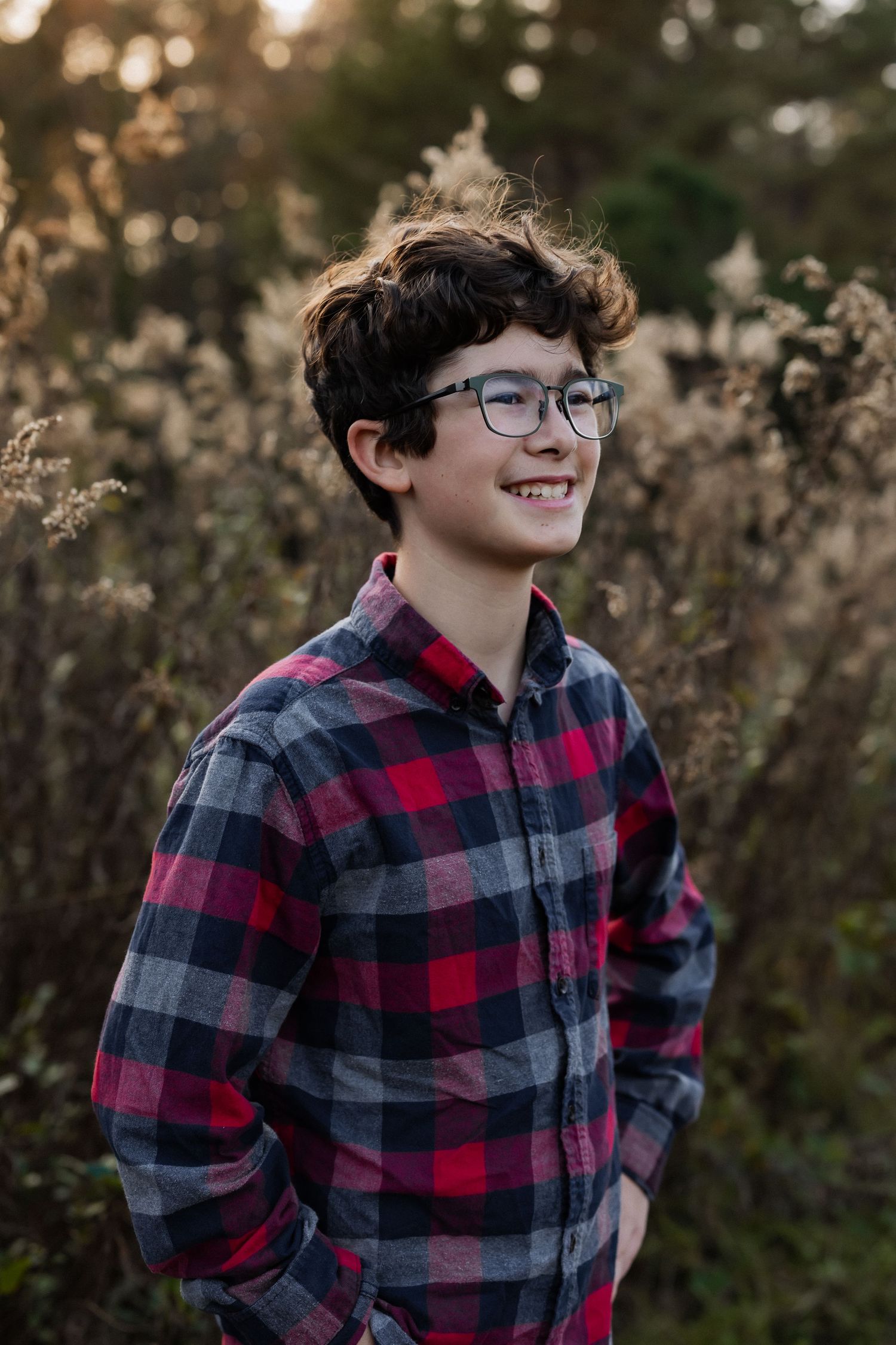 A teen in a plaid flannel shirt smiles while standing among tall grass at golden hour.