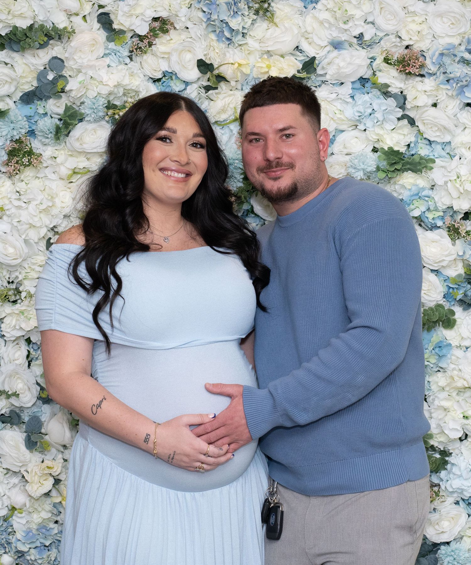 Maternity photo session with floral wall backdrop featuring couple in light blue dress and grey sweater.