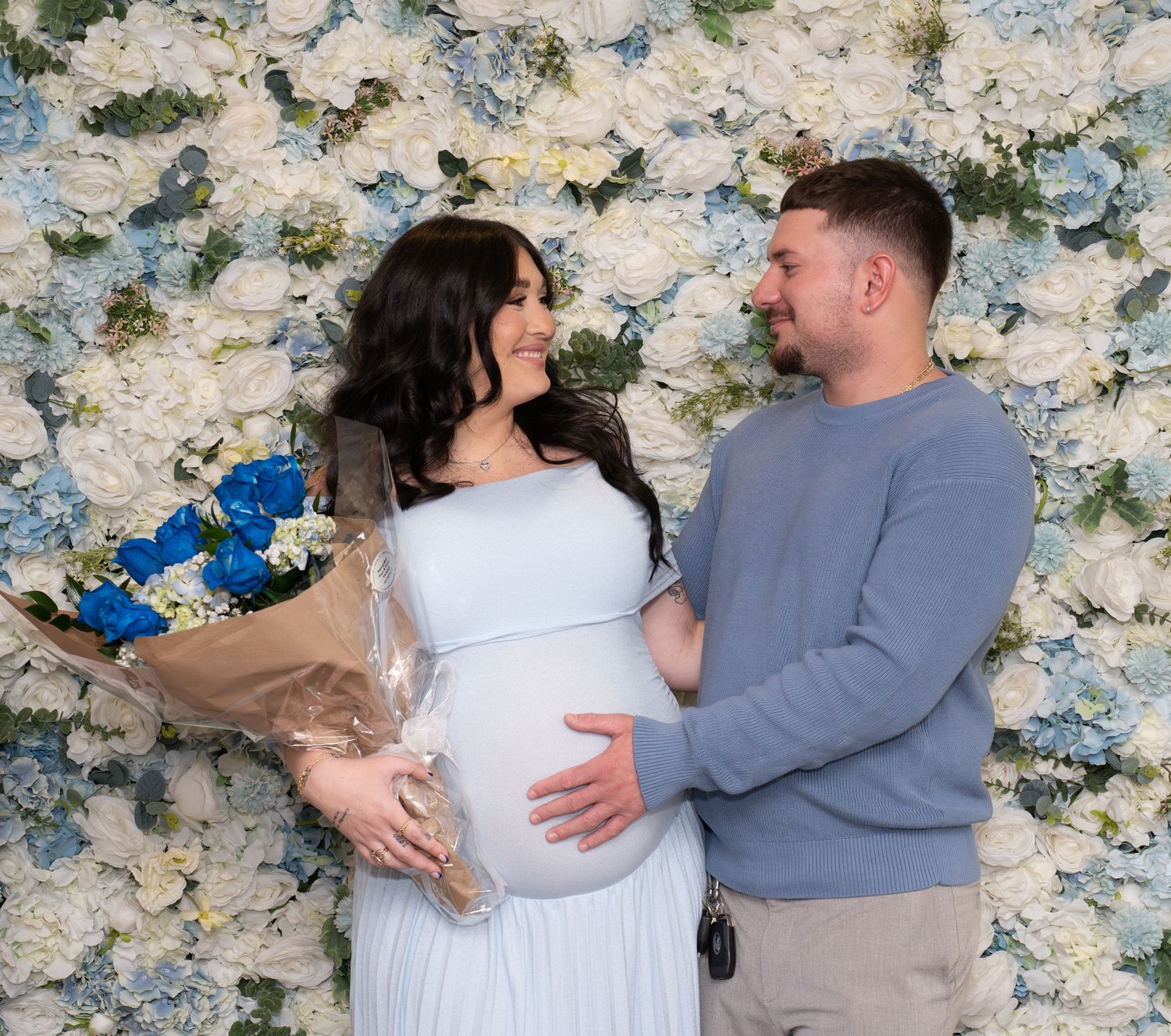 Romantic maternity photos against white floral wall with couple in coordinating blue and grey outfits.