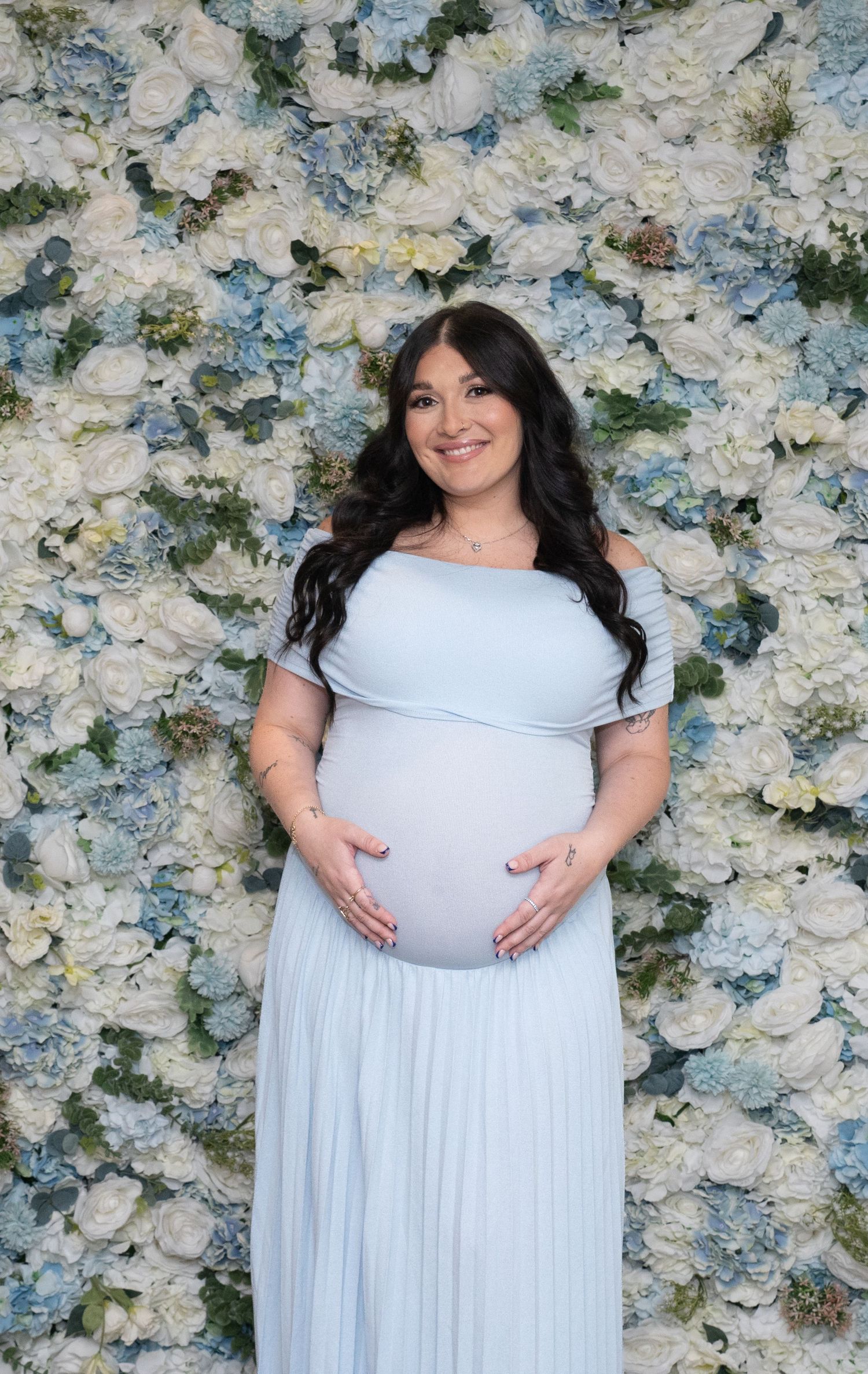 Expectant mother in flowing light blue gown poses against white floral backdrop.