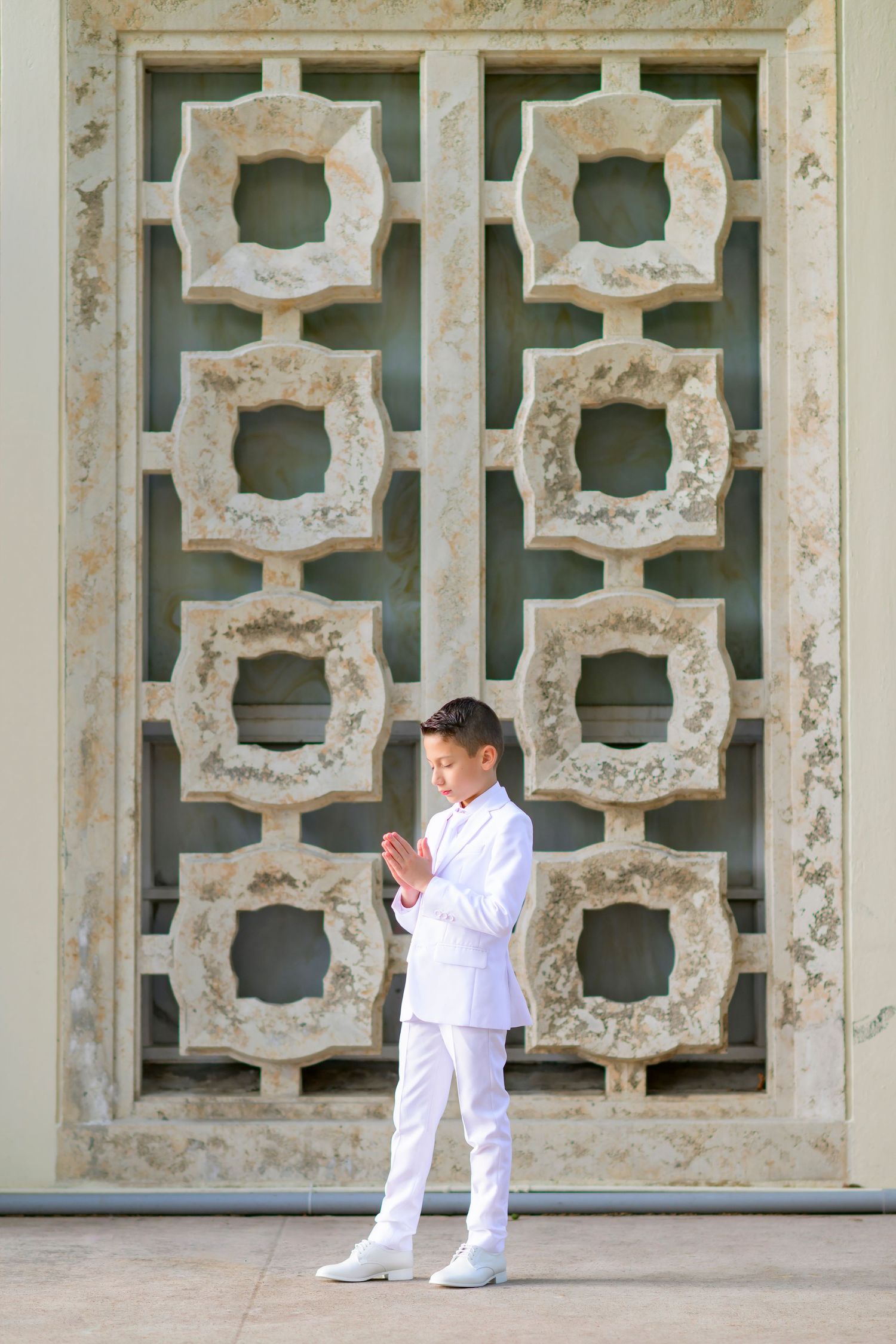 A boy in all white suit stands praying in front of ornate architectural wall with decorative geometric patterns.