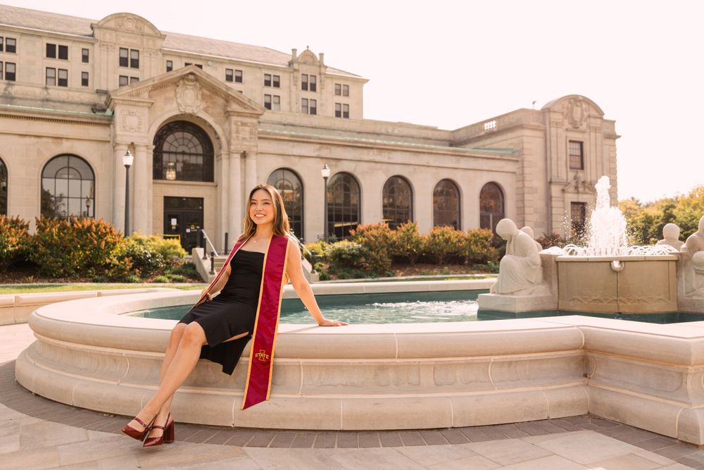 Graduate in a black dress and red stole sits on the edge of a fountain in front of a historic stone university building.