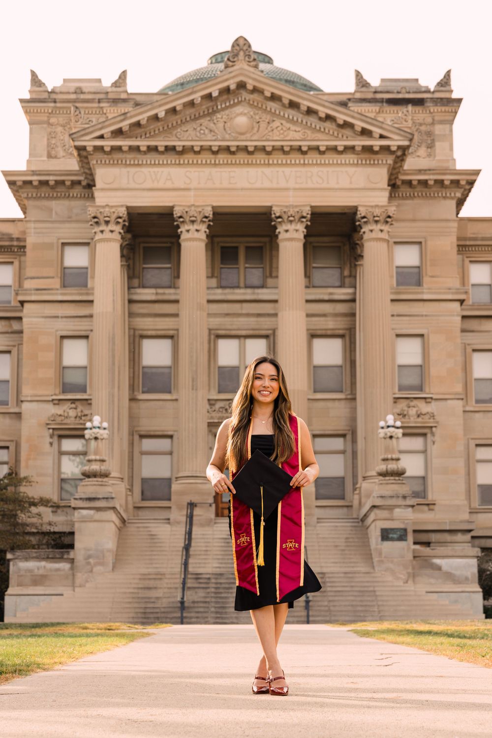 Graduate in a black dress and cardinal and gold stole holds her cap while walking in front of a historic columned university building.