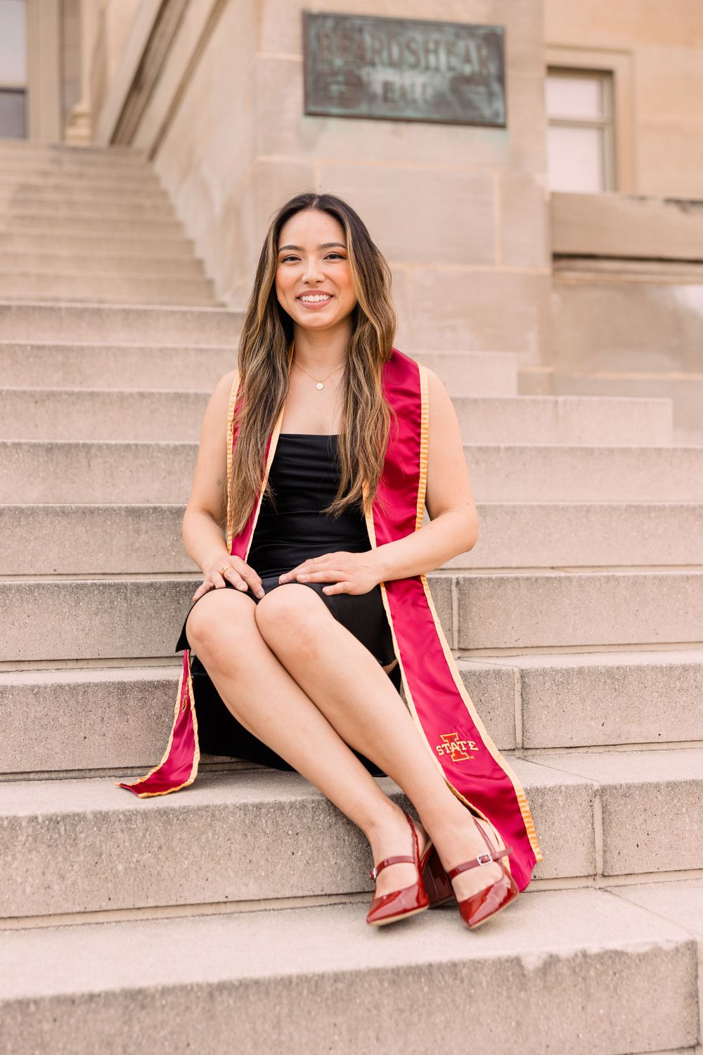Smiling graduate in a black dress and red graduation stole sits on stone steps wearing red heels outside a historic campus building.