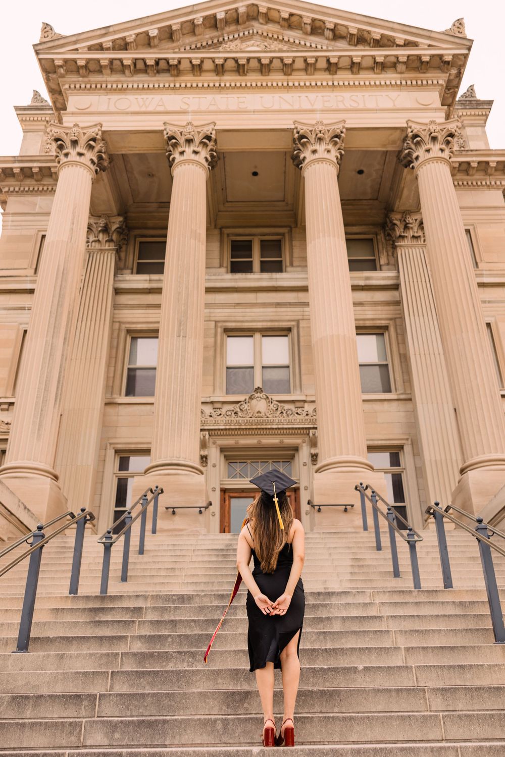 Graduate in a black cap and dress stands facing the grand columned entrance of a historic university building, seen from behind.
