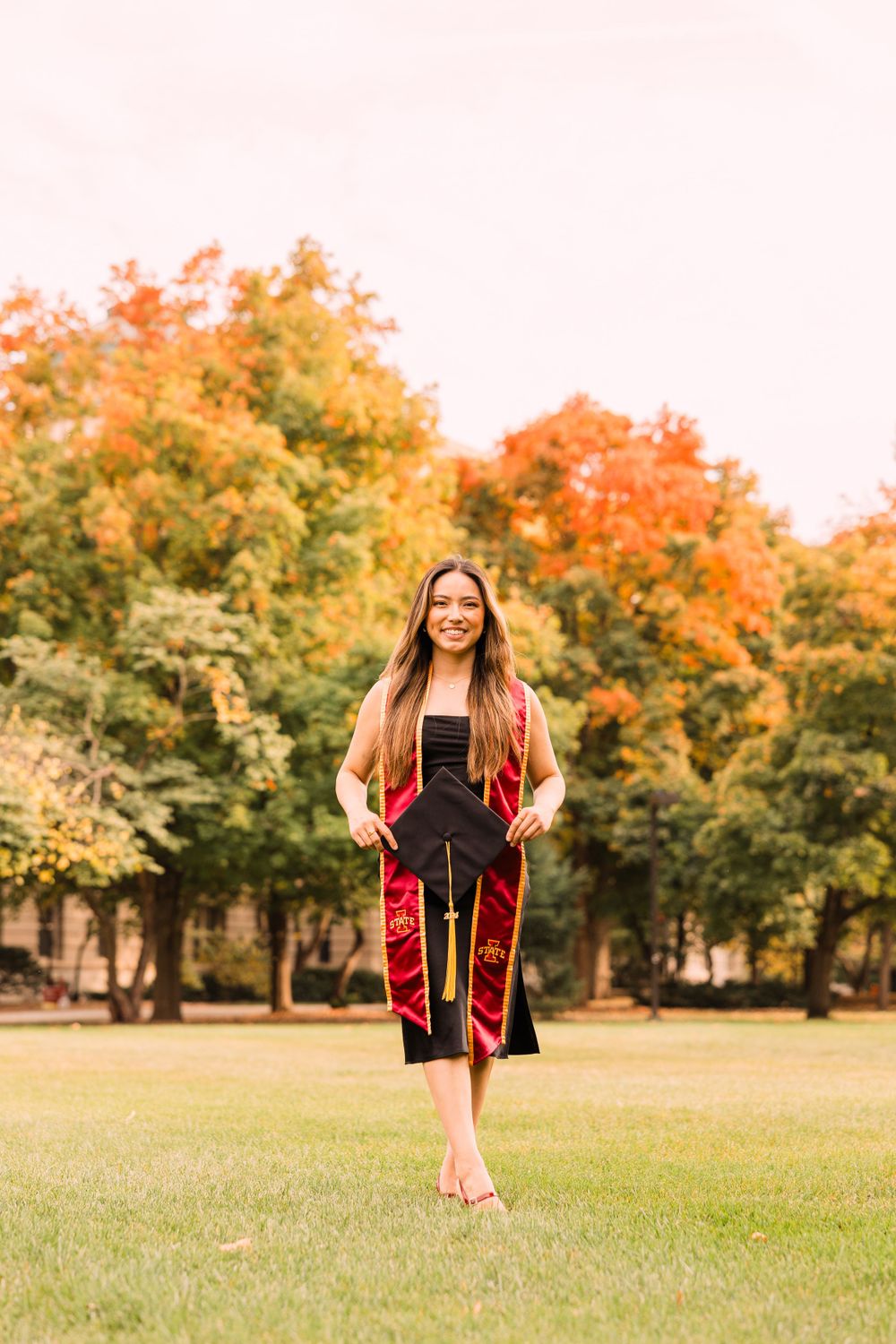 Smiling graduate in a black dress and cardinal stole carries her cap across a green campus lawn surrounded by colorful autumn trees.