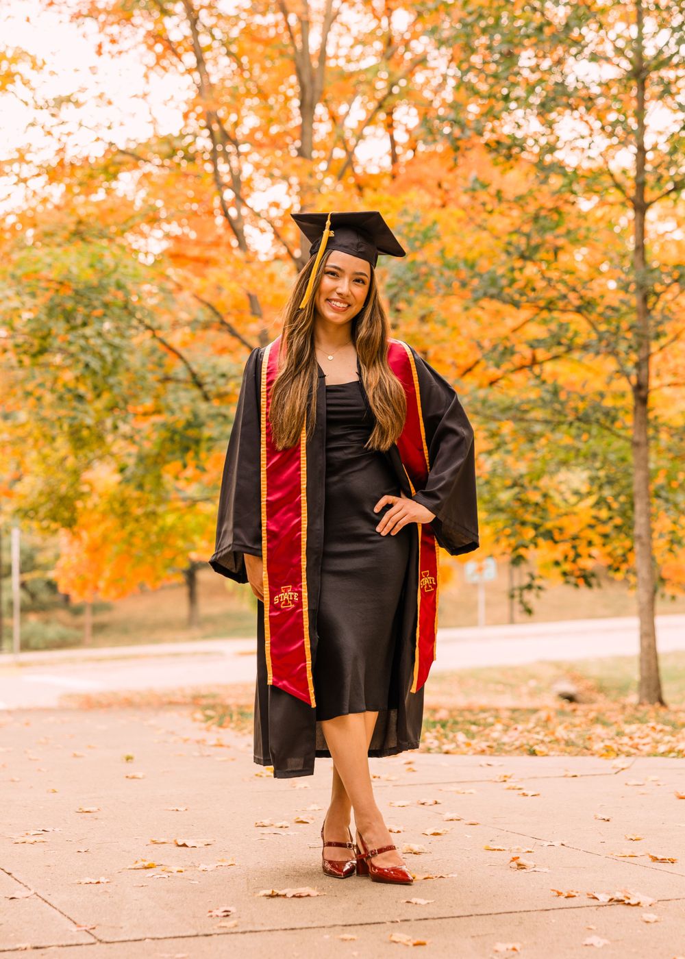 Graduate in a black cap and gown with a cardinal and gold stole poses with a hand on her hip beneath bright autumn foliage.