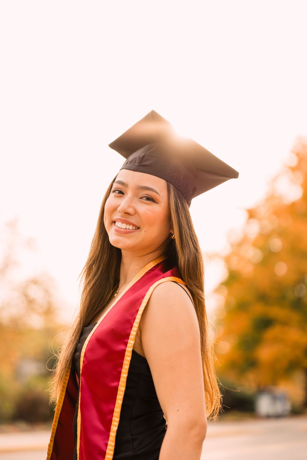 Close-up of a smiling graduate in a black cap and cardinal and gold stole with warm sunlight and autumn trees in the background.