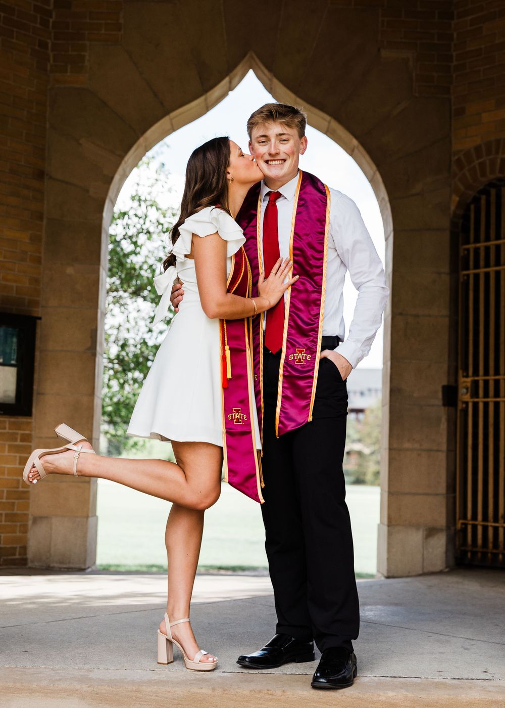 Graduating couple shares a kiss under a stone archway, she in a white dress and he in a tie, both wearing cardinal and gold stoles.