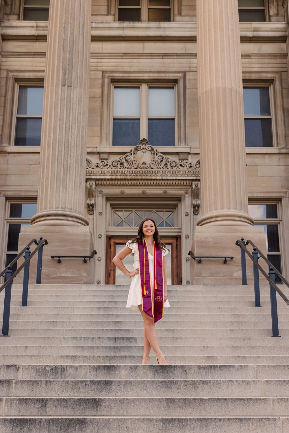 Graduate in a white dress and maroon and gold stole stands smiling on the steps of a columned historic university building.
