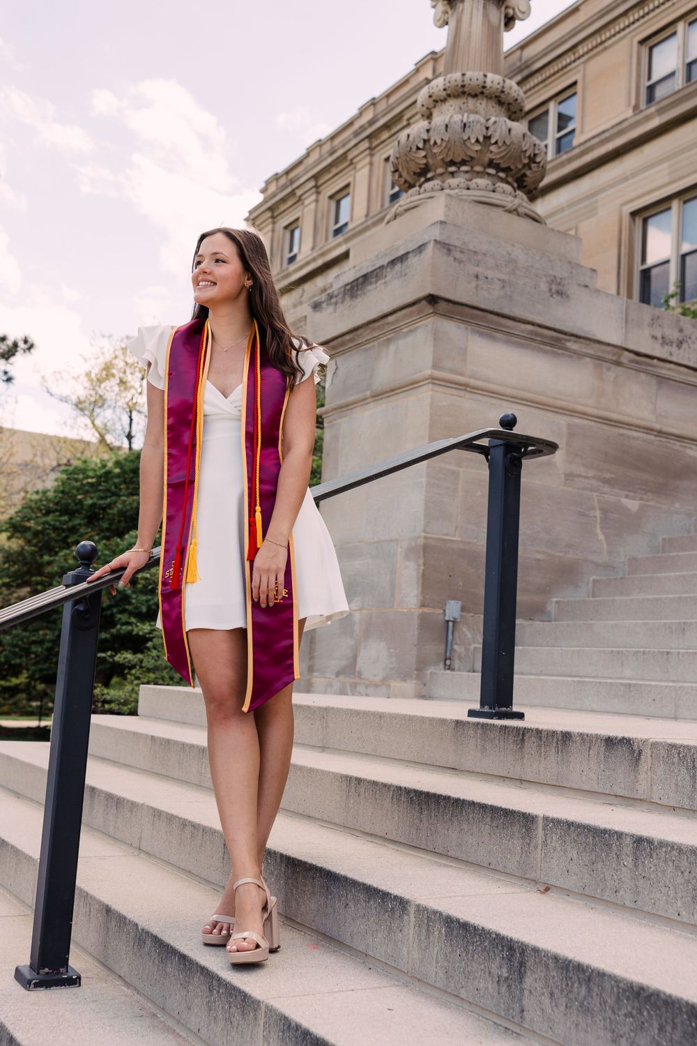 Graduate in a white dress and maroon stole smiles while standing on stone campus steps beside a black railing and ornate pillar.