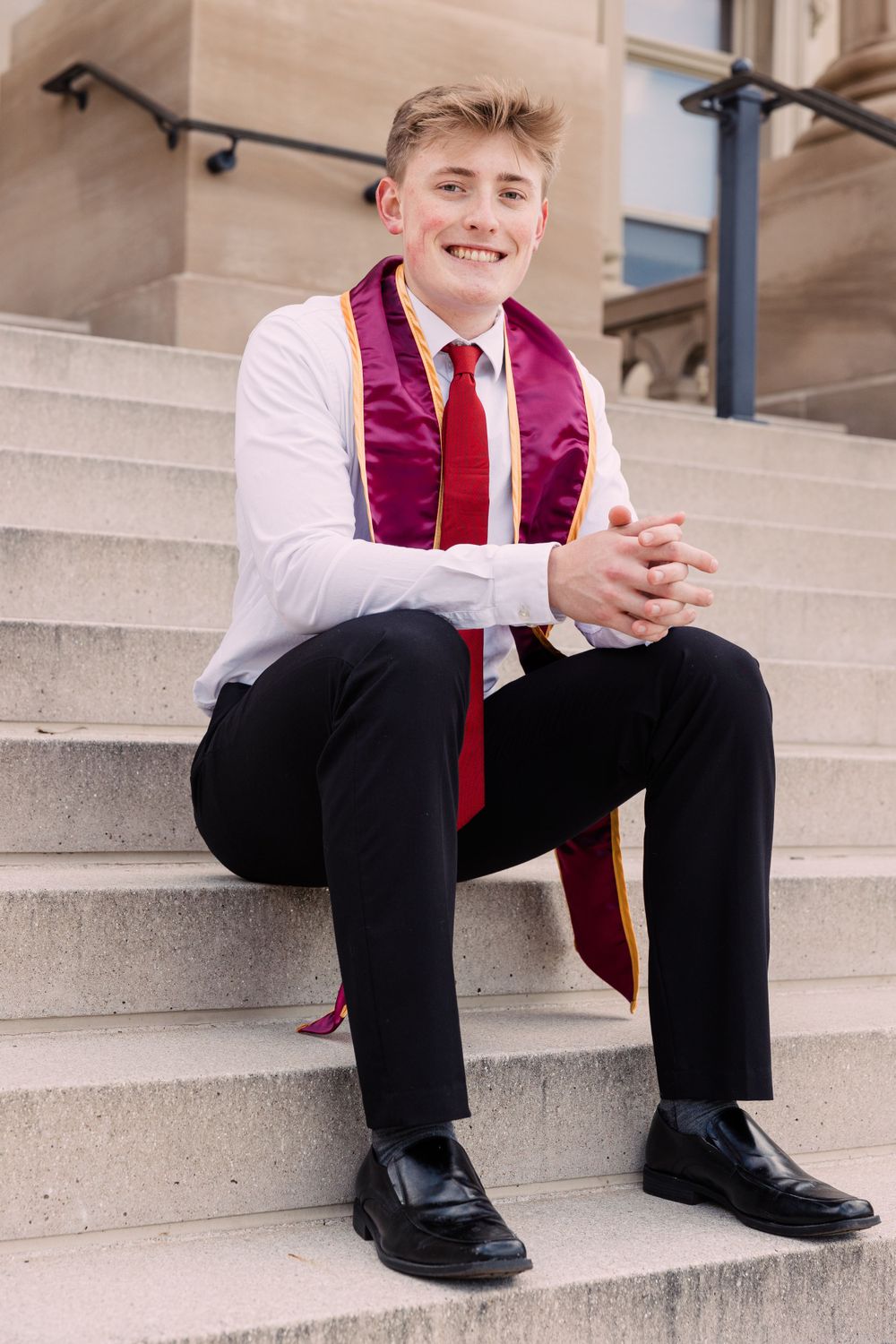 Smiling graduate in a white shirt, red tie, and maroon and gold stole sits with clasped hands on stone steps on campus.
