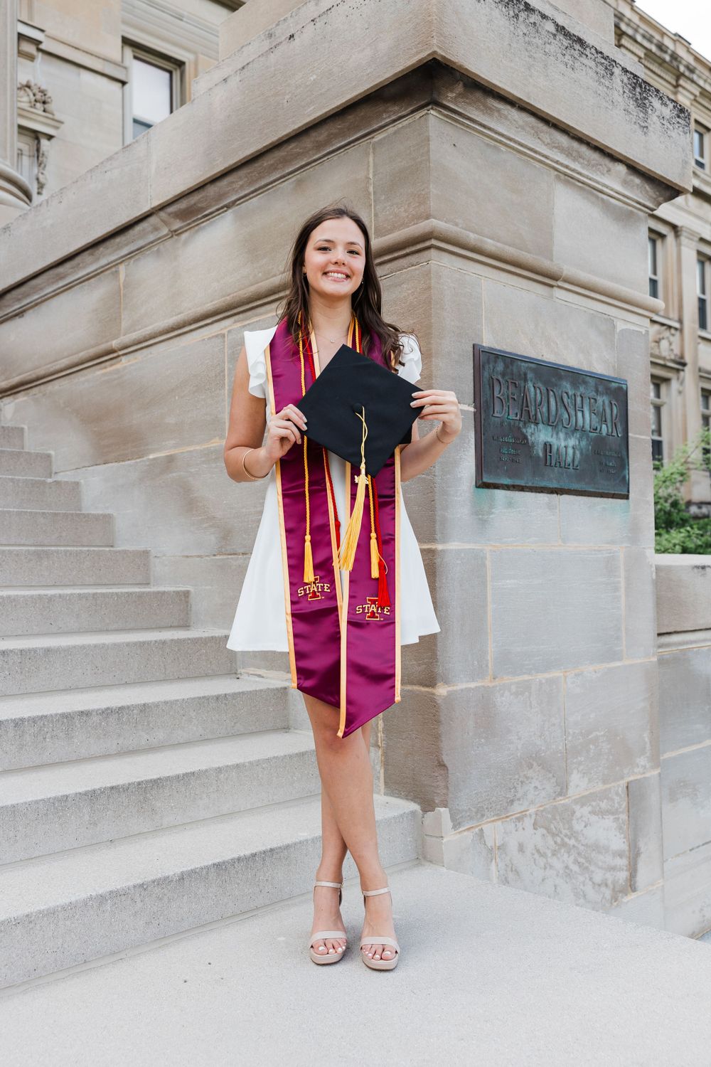 Smiling graduate in a white dress and maroon stole holds her black cap while standing beside stone steps on a college campus.