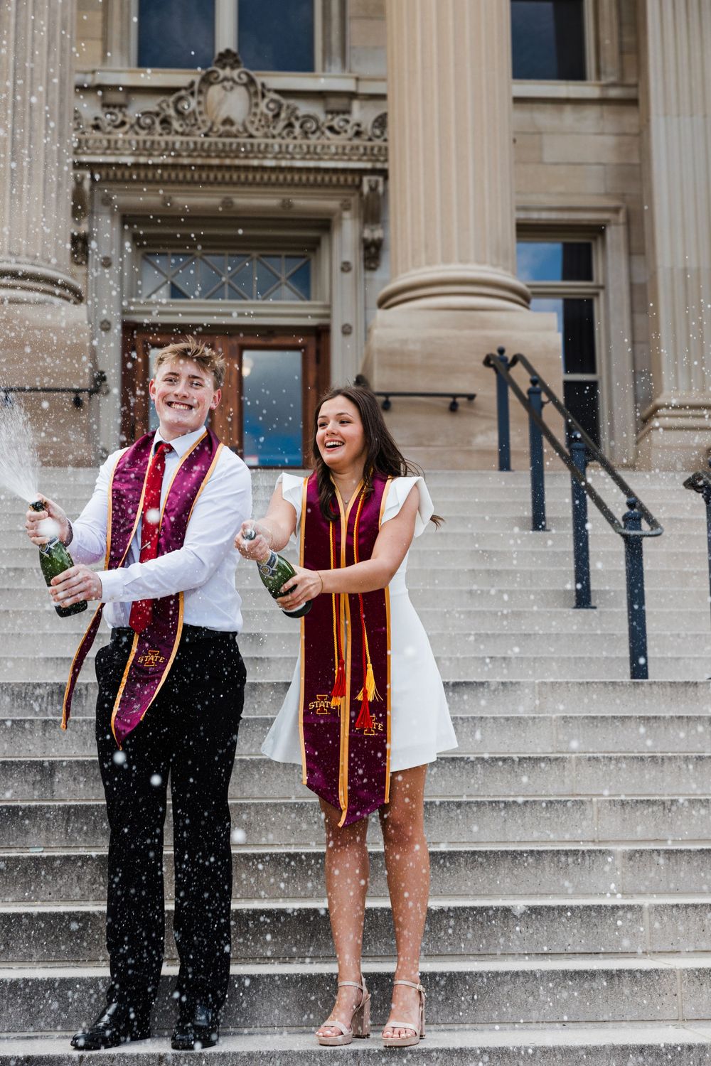 Two graduates in maroon and gold stoles pop champagne bottles and celebrate on the stone steps of a historic campus building.