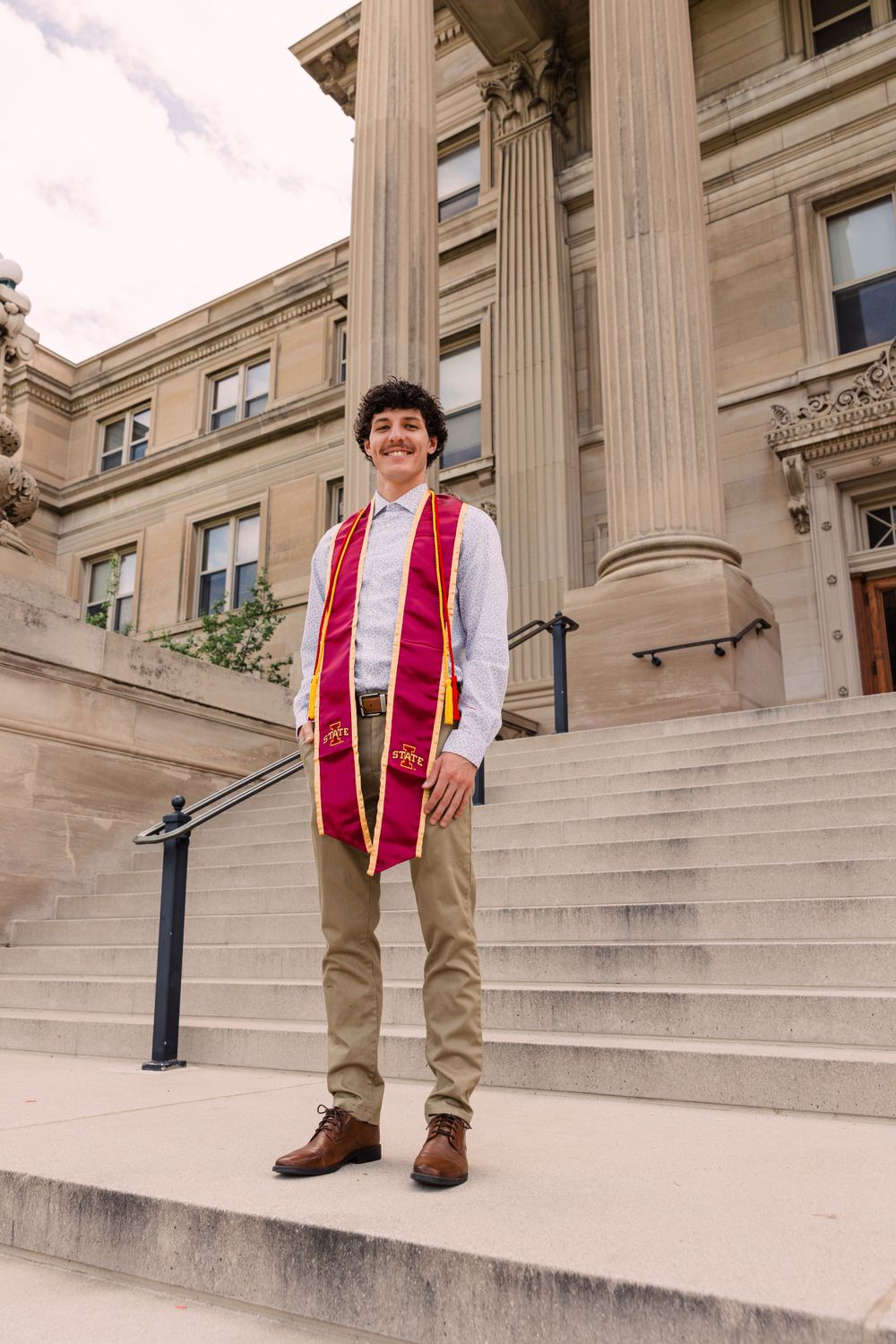 Smiling male graduate in a maroon and gold stole and khaki pants stands on stone steps in front of a columned campus building.