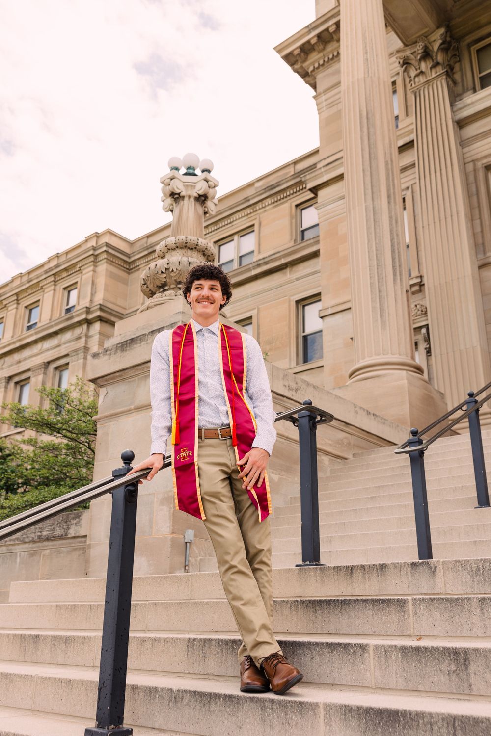 Male graduate wearing a maroon and gold stole leans casually on a black railing along stone steps beside a historic campus building.