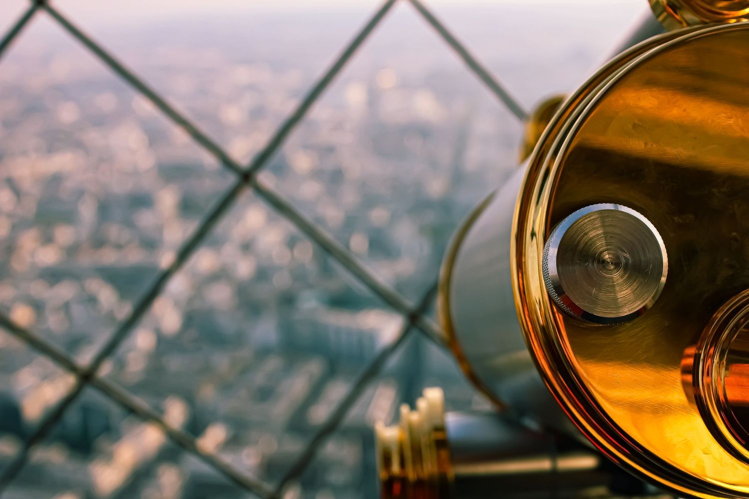 Photograph shot from top of Eiffel Tower of a golden telescope overlooking Paris through a wire barrier.