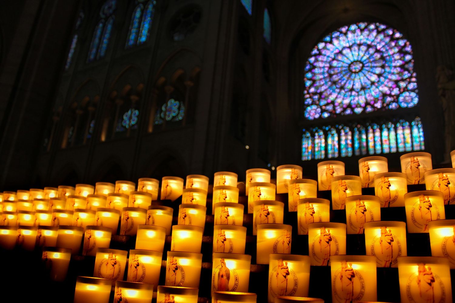 A rose window at Notre Dame cathedral in Pairs with votive prayer candles in front. Dim lighting with vibrant stained glass windows