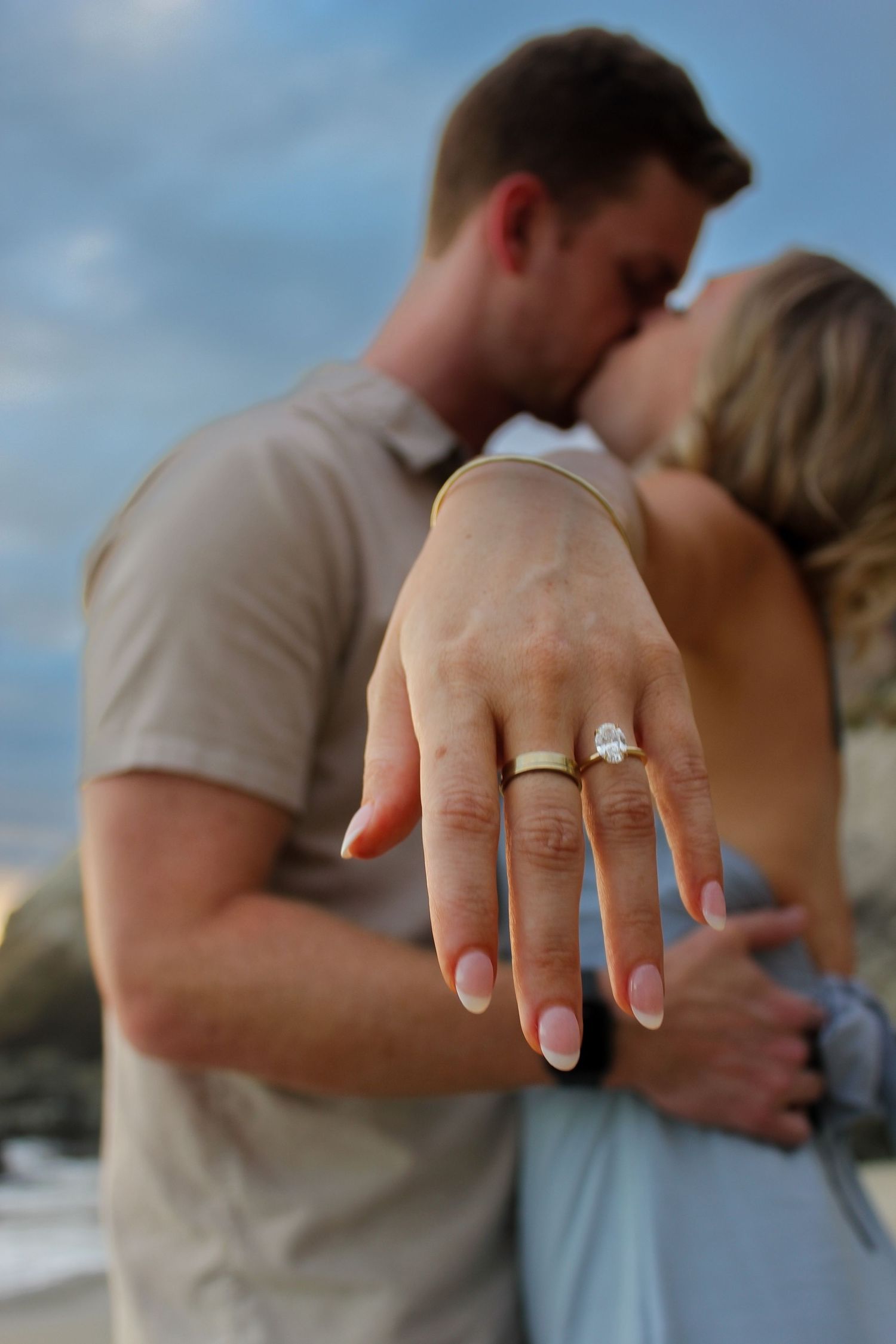 A newly engaged couple embrace each other in a kiss while the bride to be shows off her engagement ring on a beach photoshoot.
