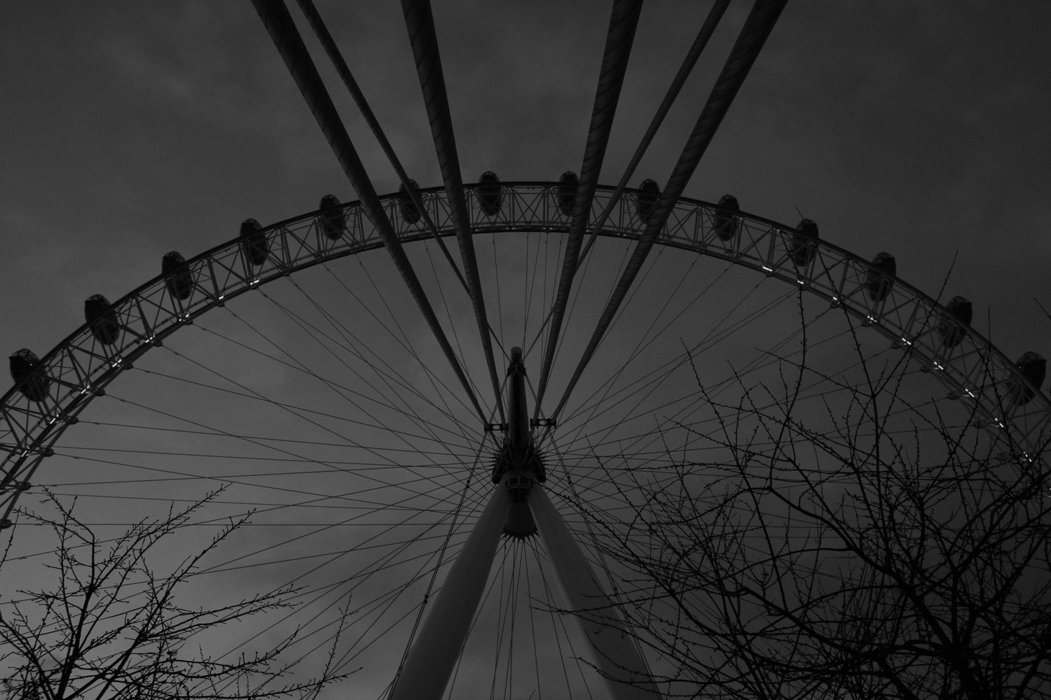 Black and white photograph looking up at the London Eye in Westminster. A moody atmosphere with trees and back supports of the ferris wheel