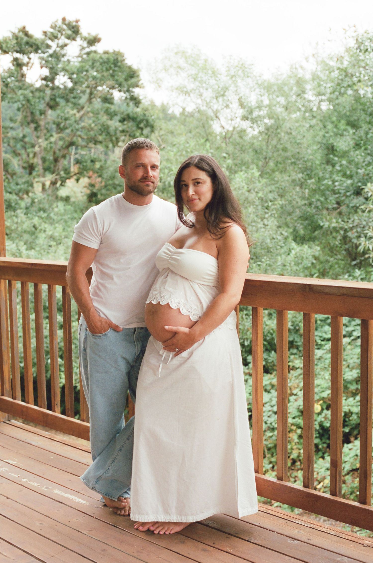 Couple poses on wooden deck, pregnant woman in white dress cradling baby bump, man in white tee and jeans, lush greenery background. Film