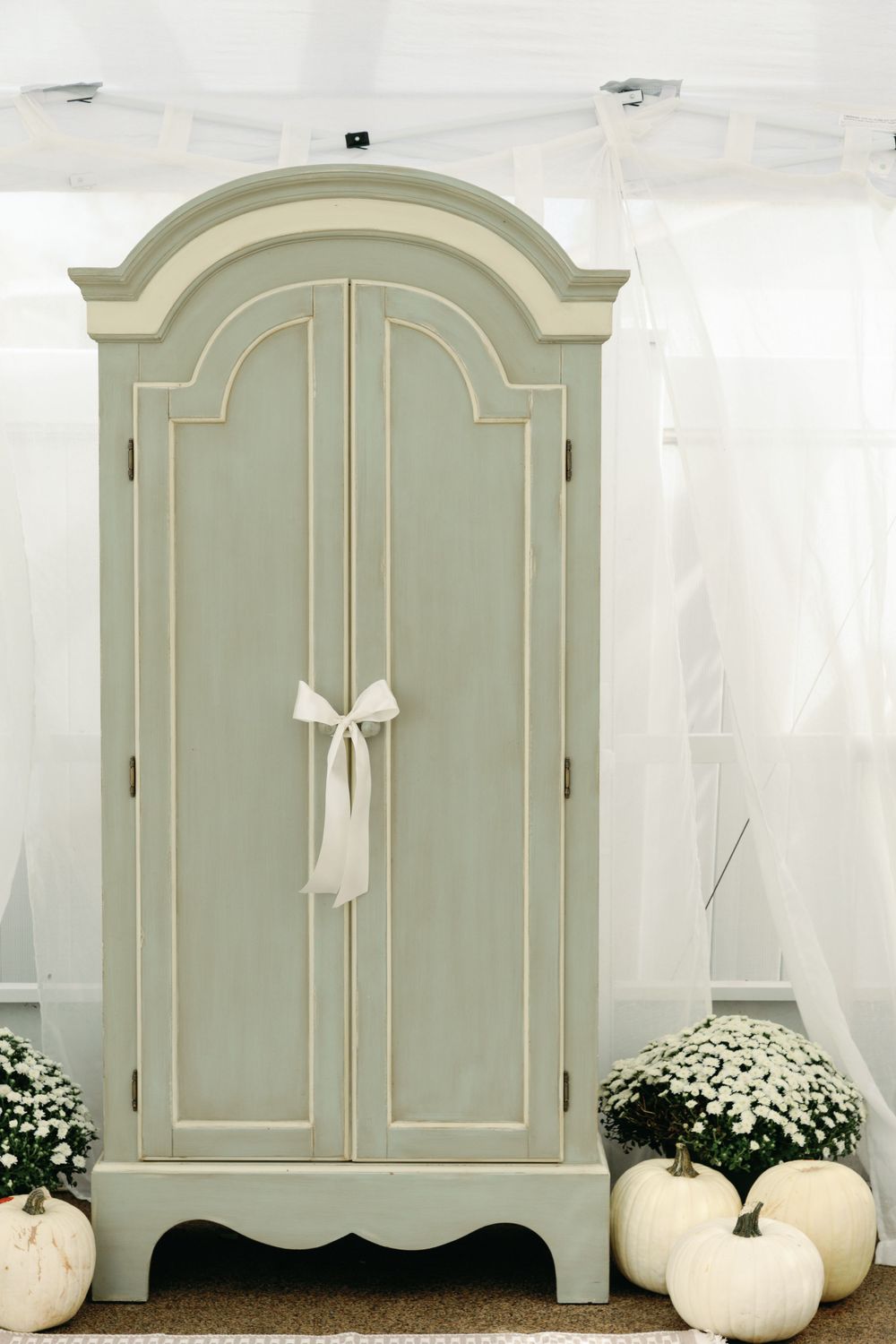 Elegant sage green armoire with white bow, surrounded by white pumpkins and chrysanthemums.