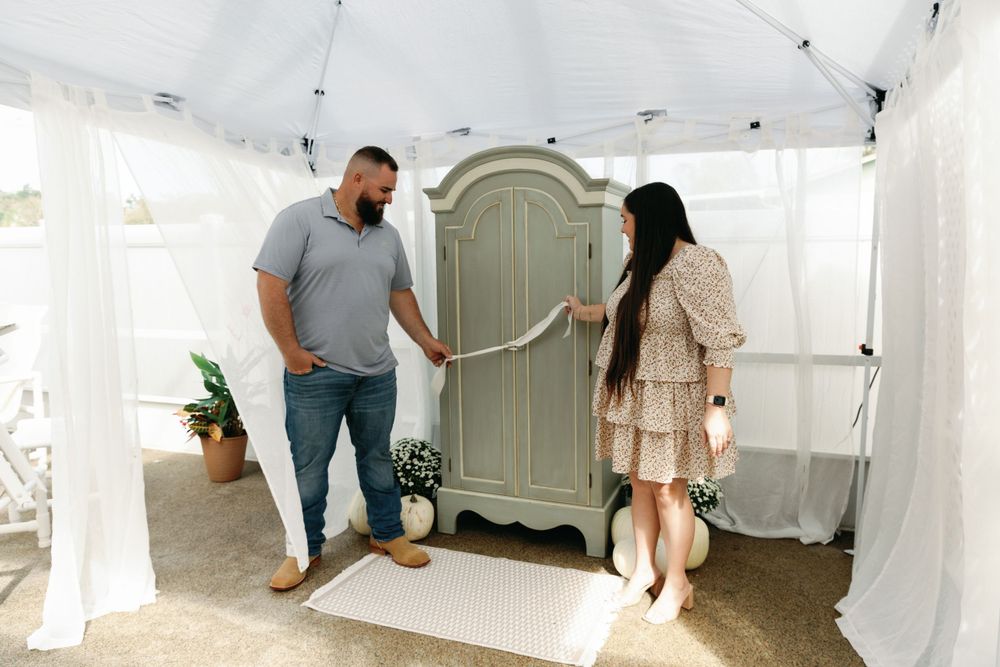 Couple standing beside closed sage green armoire at an outdoor gender reveal party.