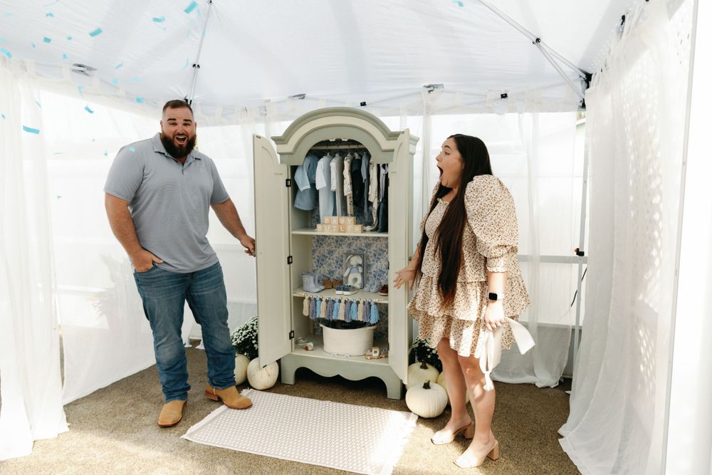 Couple smiling beside open sage green armoire filled with baby boy clothes at gender reveal.