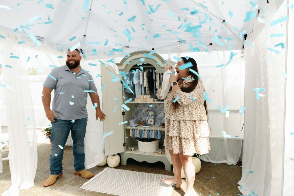 Couple celebrates baby gender reveal with blue confetti falling under a white tent.