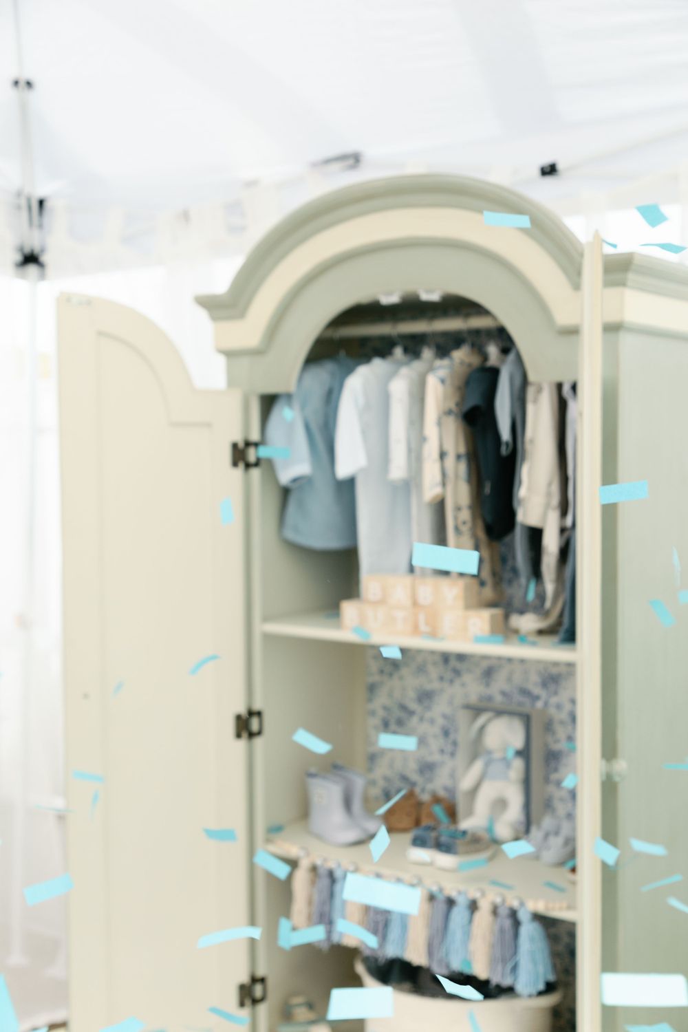 Blurred sage armoire filled with baby clothes and blue confetti during gender reveal.