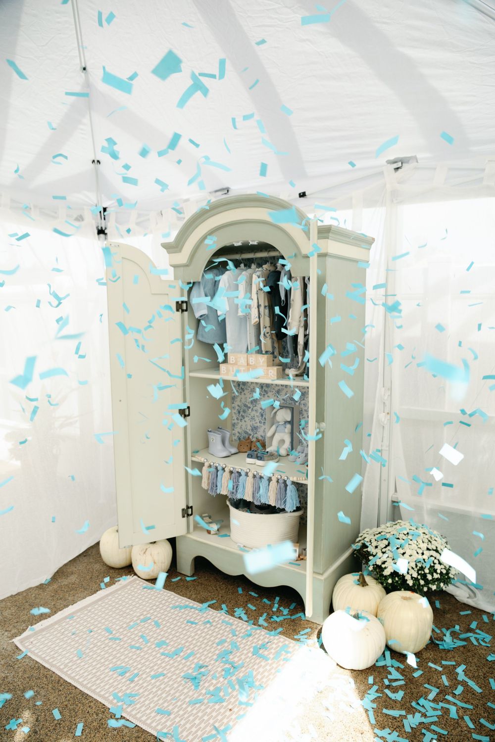 Blue confetti fills white tent around sage armoire during baby boy gender reveal.