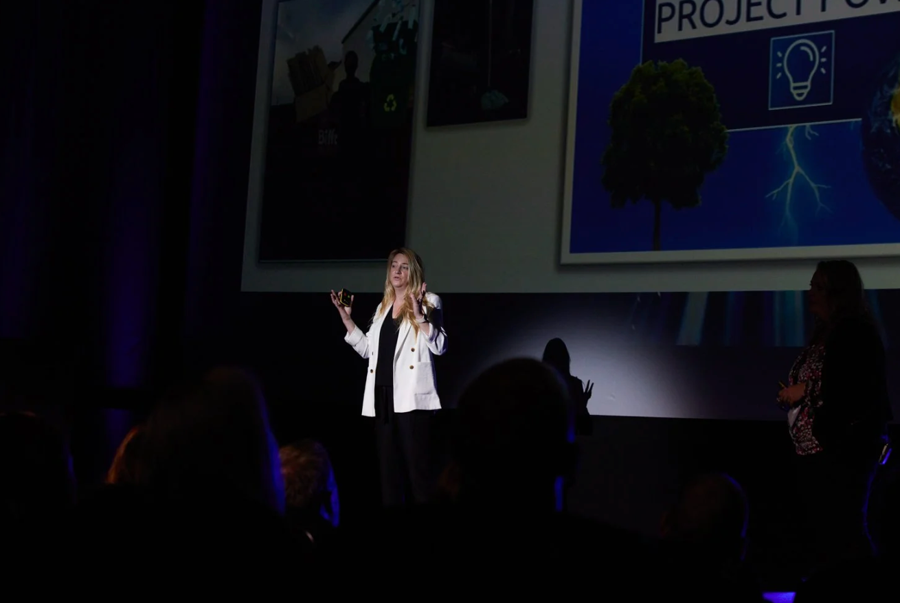A person in a white lab coat gestures while speaking on stage during a presentation.