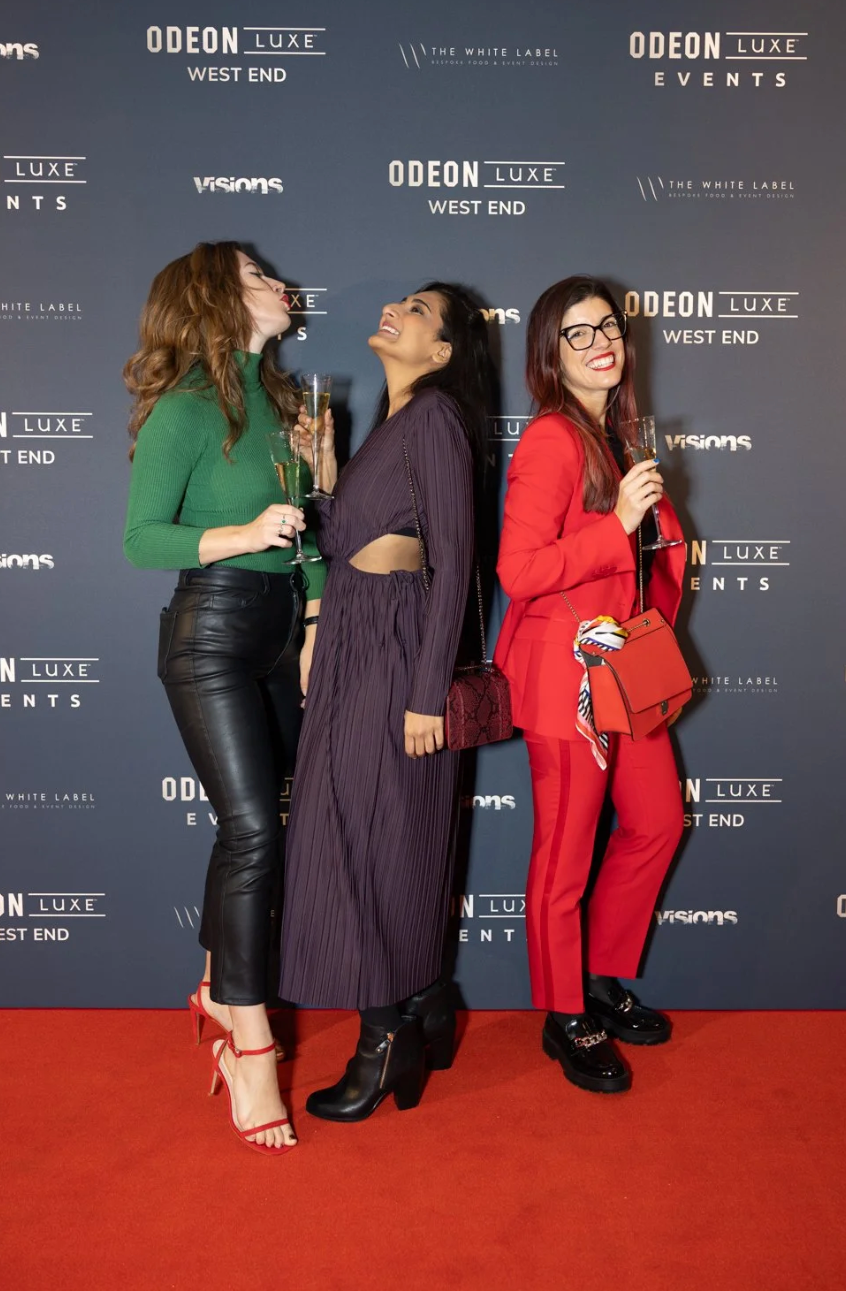 Three friends in stylish outfits laugh together while posing on a red carpet at an Odeon Luxe event.