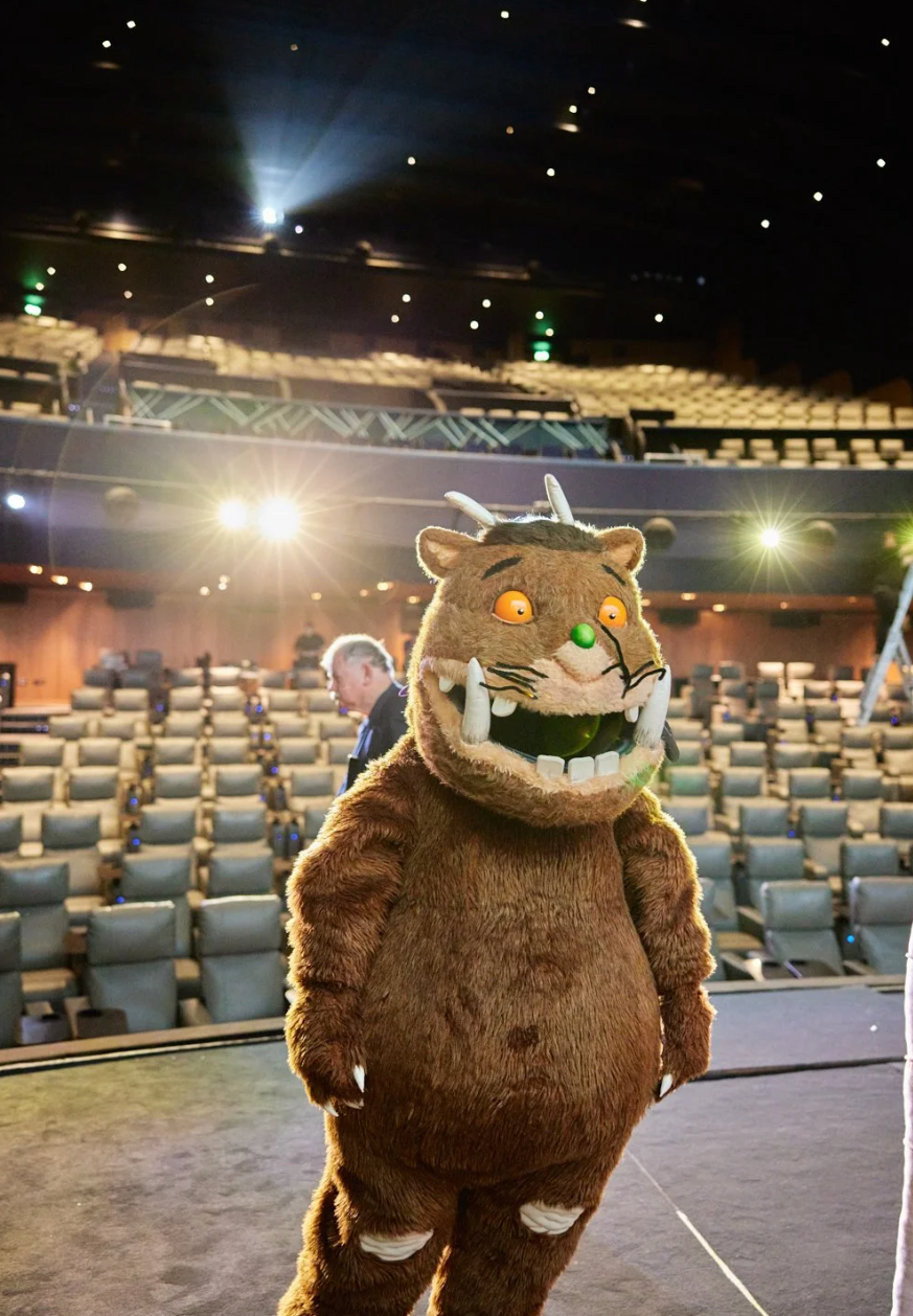 A mascot in a brown furry costume performs on an empty stage with theater seats visible in the background.
