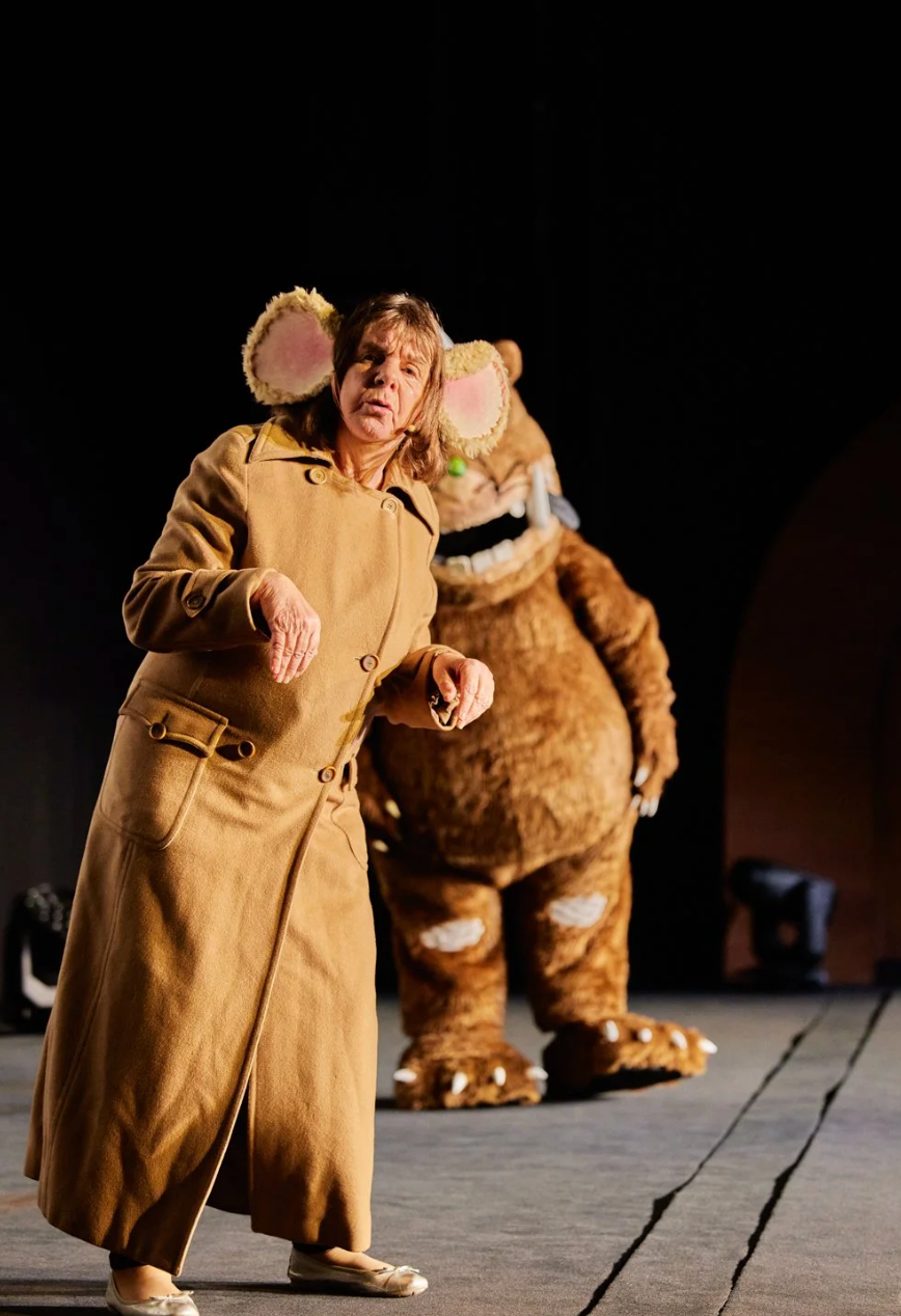 A person in a tan coat performs on stage with a large brown monster costume in the background.