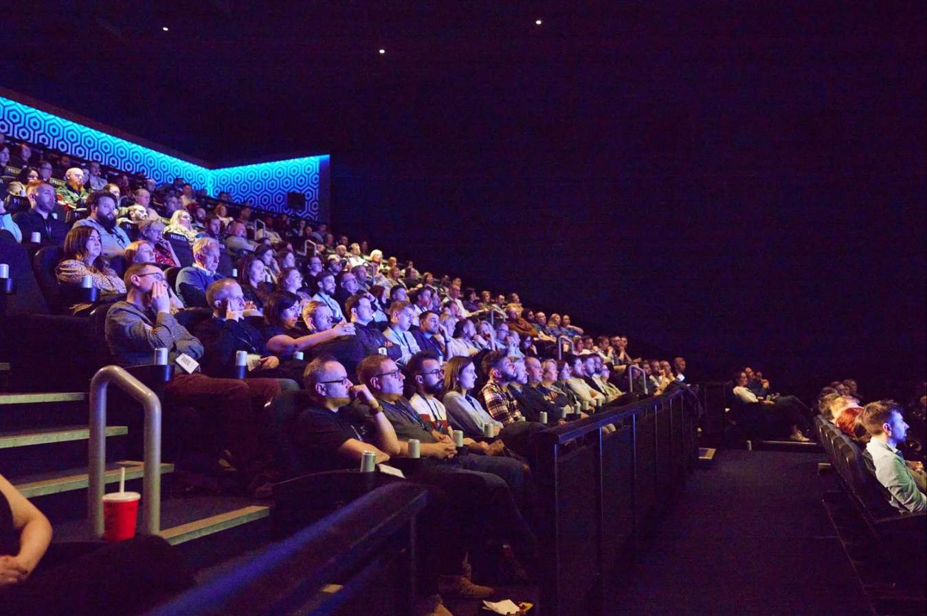 Large audience seated in tiered theater seating with blue lighting during a presentation or performance.