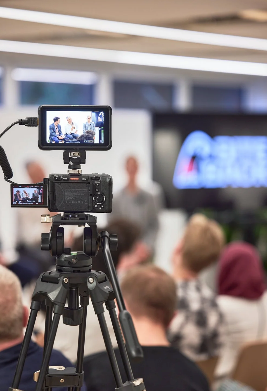 Video camera mounted on tripod recording audience at indoor conference event.
